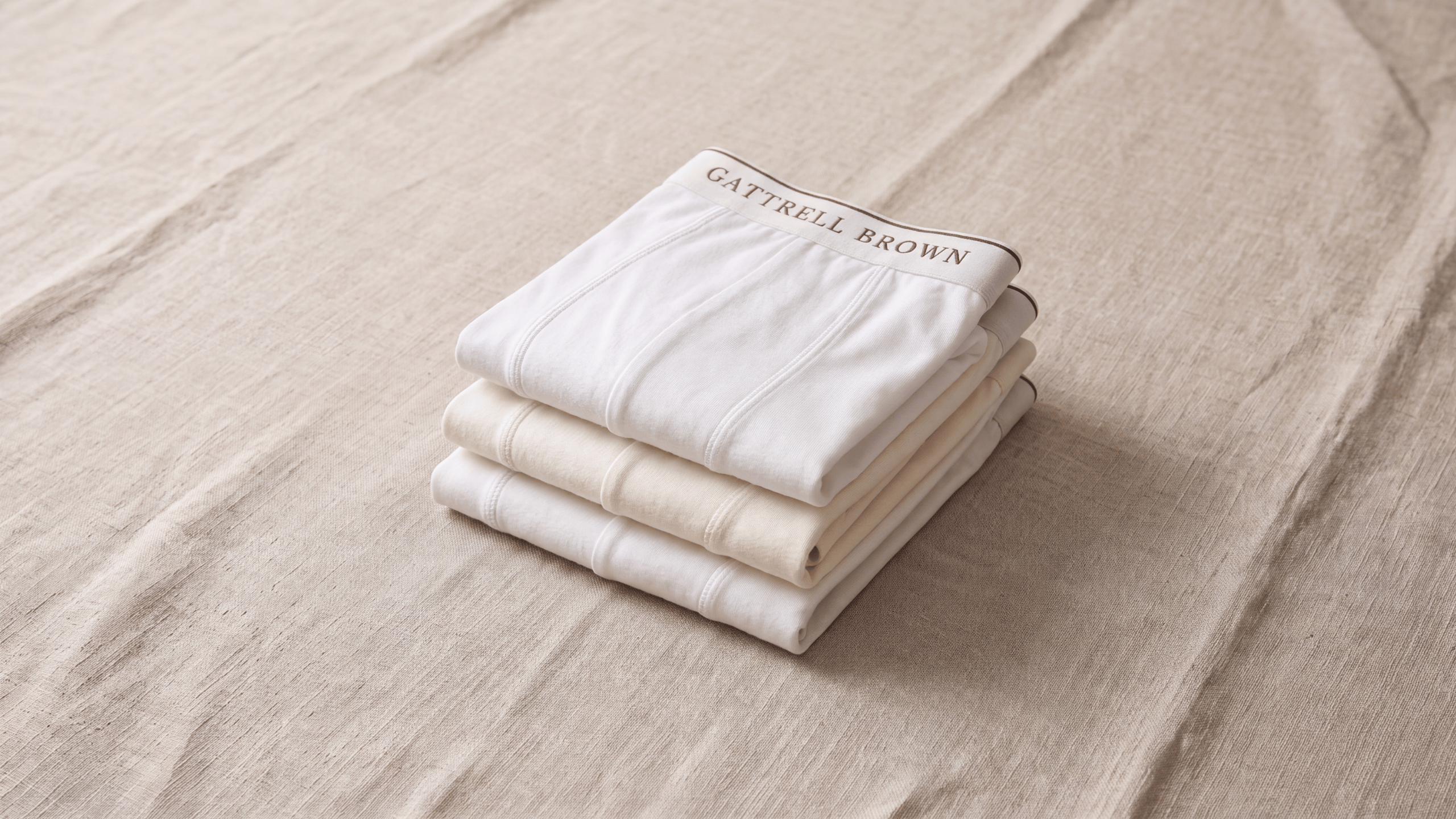 Three folded white and cream bedding pieces stacked on a beige linen surface, with the top piece showing the brand name Gattrell Brown.