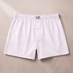 White ribbed cotton boxer briefs with a button fly and an elastic waistband featuring a brown 'GB' monogram logo, laid flat on a textured beige background.