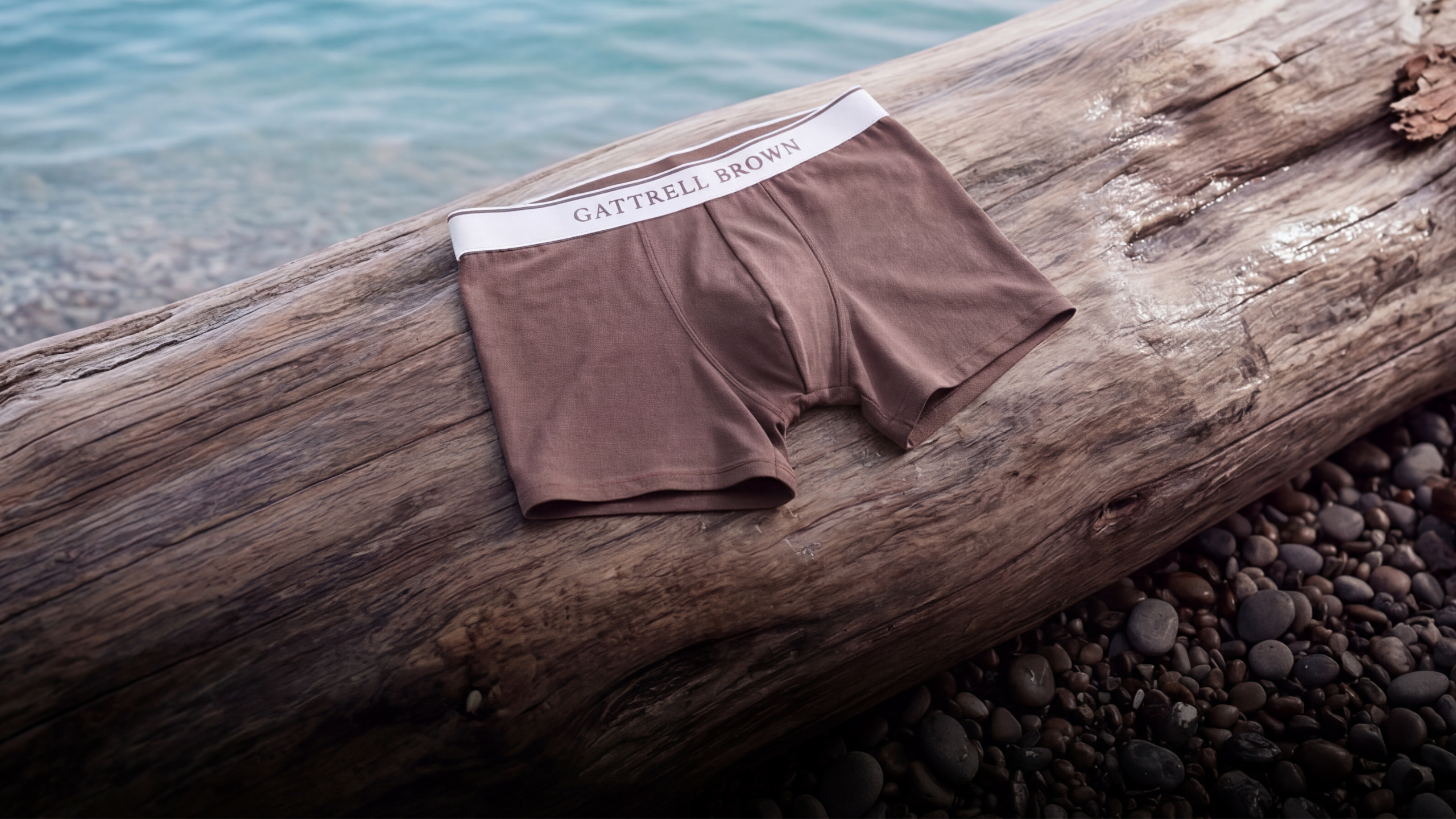 A pair of brown boxer briefs with a white elastic waistband featuring the brand name 'GATTRELL BROWN', laid flat on a large weathered log on a pebble beach with water in the background.